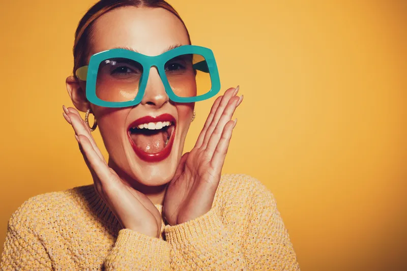 Excited woman with large blue sunglasses and red lipstick smiles widely, wearing a yellow sweater against an orange background. Concept of joy, fashion, and vibrant style.