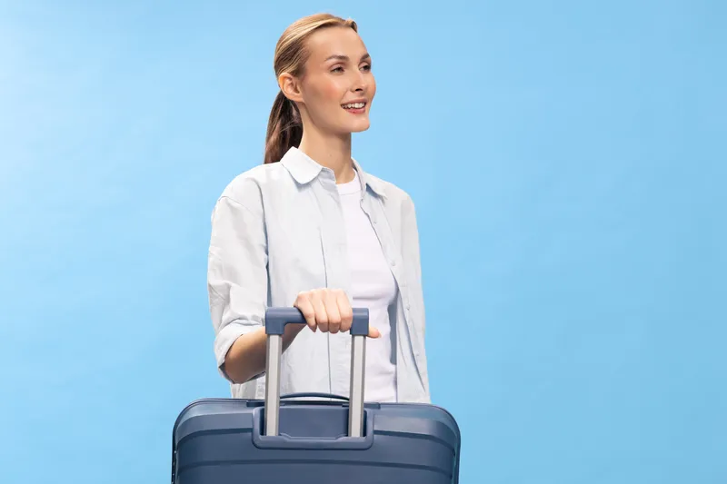 Young woman with suitcase stands smiling against blue background. She is ready for travel, wearing casual white shirt and looking away with positive mood.