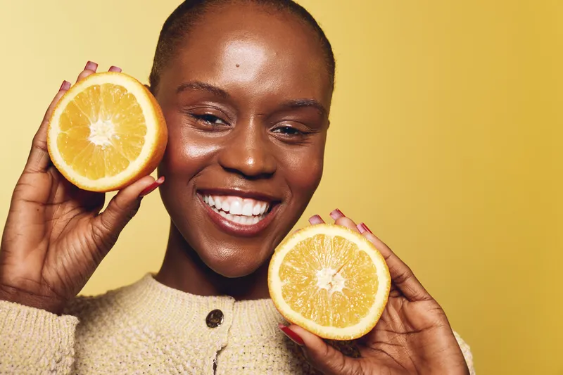 Smiling African woman with dark skin holding two halves of fresh orange near face on yellow background. Concept of healthy lifestyle, natural beauty, vitamin C, and skincare wellness.