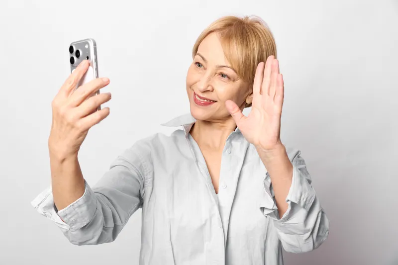 Woman elderly selfie smartphone smiling touch cheek skincare concept, holding smartphone for selfie, smiling and touching cheek in studio, selfcare beauty routine, soft light on white background