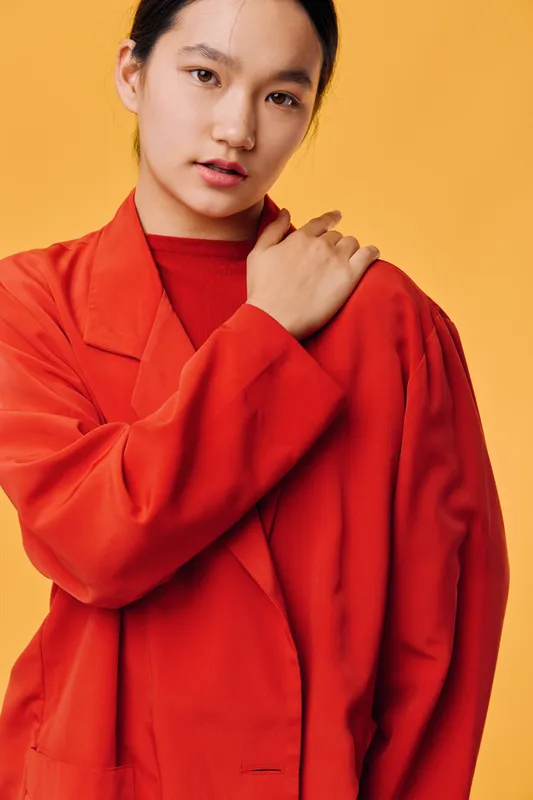 Portrait of young woman in red coat posing on yellow background with hand on shoulder, looking at camera with confident facial expression and neat hairstyle in studio light.