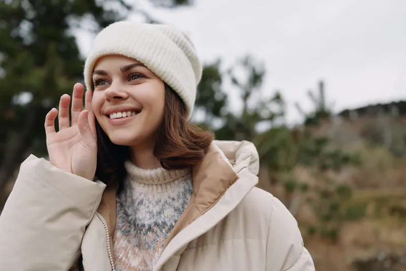 Woman outdoors wearing a warm coat and white beanie smiling and waving hello, natural landscape with trees and soft daylight, casual lifestyle moment