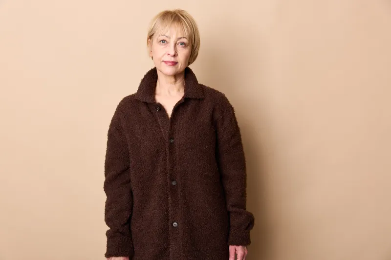 Woman portrait in studio, mature woman wearing winter coat, beige background, calm smile, standing indoors, soft lighting, warm outerwear texture, framing