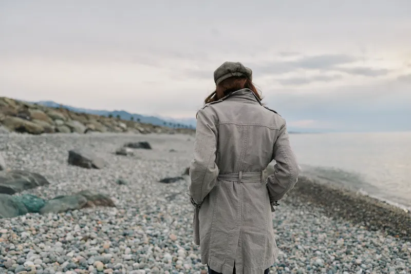 Woman in warm coat and hat stands alone on rocky beach facing sea under cloudy sky. Concept of solitude, cold weather, and peaceful outdoor scenery near water.