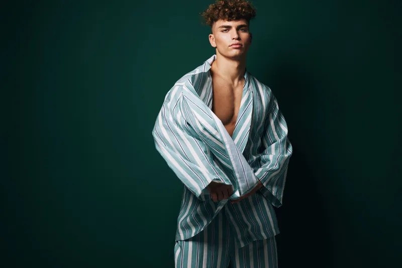 Young man with curly hair wearing striped oversized shirt and pants posing against dark background with serious expression and confident posture in studio lighting.