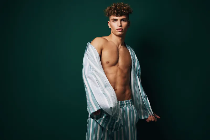 Young man with curly hair wearing striped pajamas posing shirtless against dark background, showing toned body and confident expression in studio lighting.