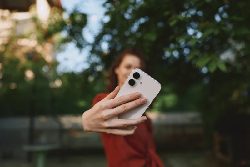 Selfie of a smiling woman in a red top taking a smartphone photo outdoors, lush trees in background, natural light, candid lifestyle moment