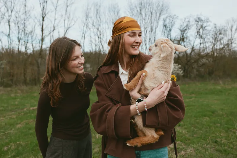 Two women outdoors share a joyful moment with a playful goat kid, creating a warm lifestyle scene of friendship, kindness, and rural charm in daylight.