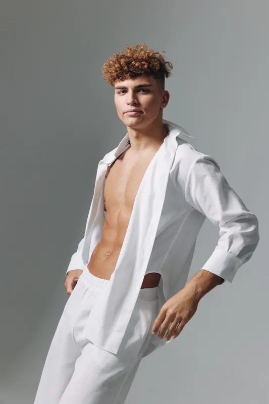 Young man with curly hair posing in white open shirt and pants against gray background. Casual fashion, confident model with toned body and relaxed expression in studio lighting.