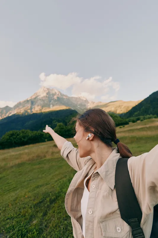 Young woman with brown hair and flower in ear points excitedly towards a mountain peak while standing in a green meadow under a clear sky.