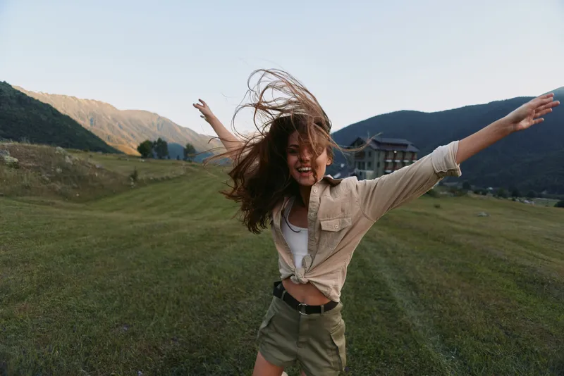 Young woman with long brown hair joyfully spins with arms outstretched in a green mountain meadow during late afternoon, expressing freedom and happiness.