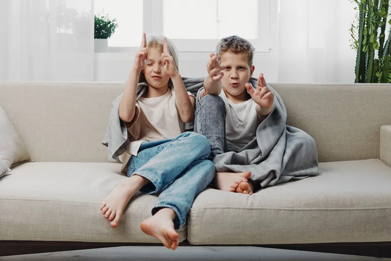 Two children sitting barefoot on a sofa wrapped in blankets playfully gesture with their hands in a bright living room setting with plants.