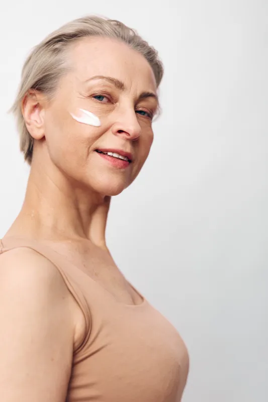Mature woman with short hair applies moisturizing cream on cheek, smiling softly. Concept of skincare, aging, self-care, beauty routine, and natural skin health in minimal studio setting.
