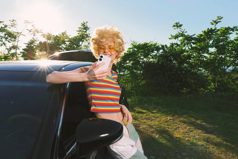 Smiling woman in a striped top leans on a car, taking a selfie with a smartphone. Outdoor scene with bright sun, retro film color, and casual fashion mood, under warm golden light and a breezy