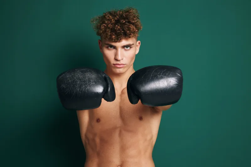 Young man with curly hair wearing black boxing gloves in fighting stance against green background. Shirtless athlete training for boxing or martial arts with focused expression.