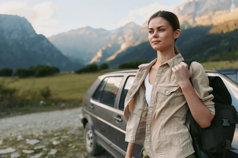 Confident woman outdoors, standing beside a car with a backpack, ready for a hike, rugged mountains and open valley behind, warm light and fresh air.
