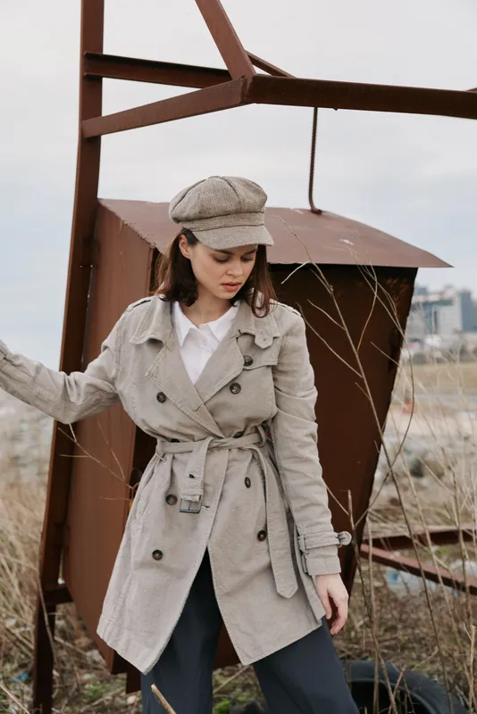 Young woman in a beige trench coat and matching hat stands near rusty metal structure outdoors. Overcast sky and dry grass create a moody urban exploration atmosphere.
