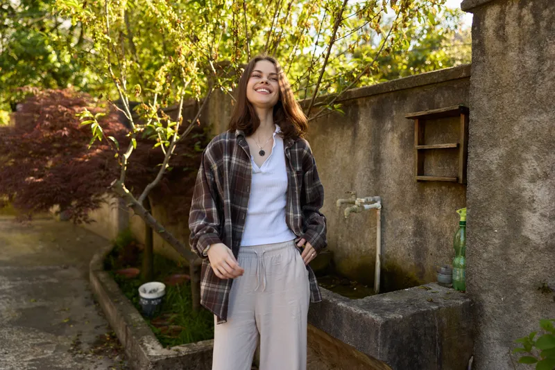 Smiling woman in casual clothing stands outdoors in sunlight, embracing a relaxed lifestyle. A bright garden setting frames a cheerful, everyday moment