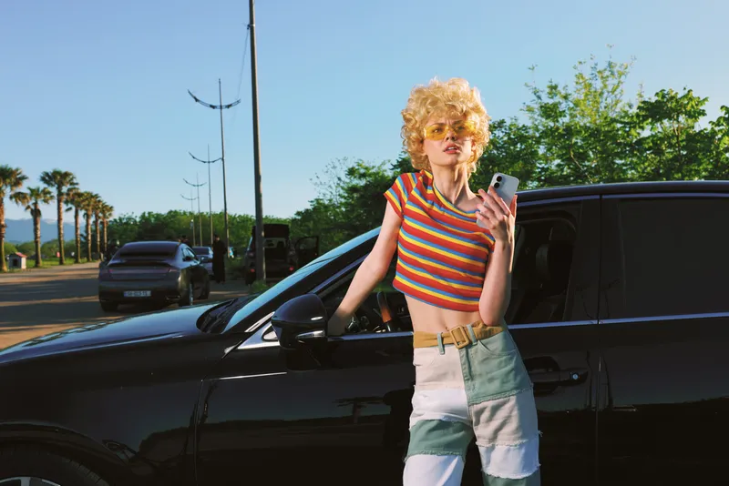 A stylish woman in a striped crop top and patchwork shorts leans on a dark car, checking her phone under bright sun. Retro film color tones create a nostalgic, carefree city lifestyle vibe.