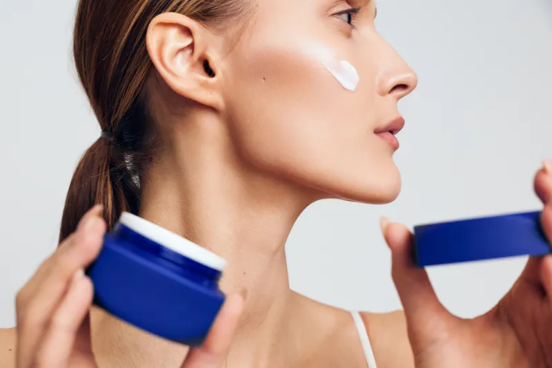 A young woman applies facial cream from a blue jar in profile, showcasing a skincare routine and moments of self care.