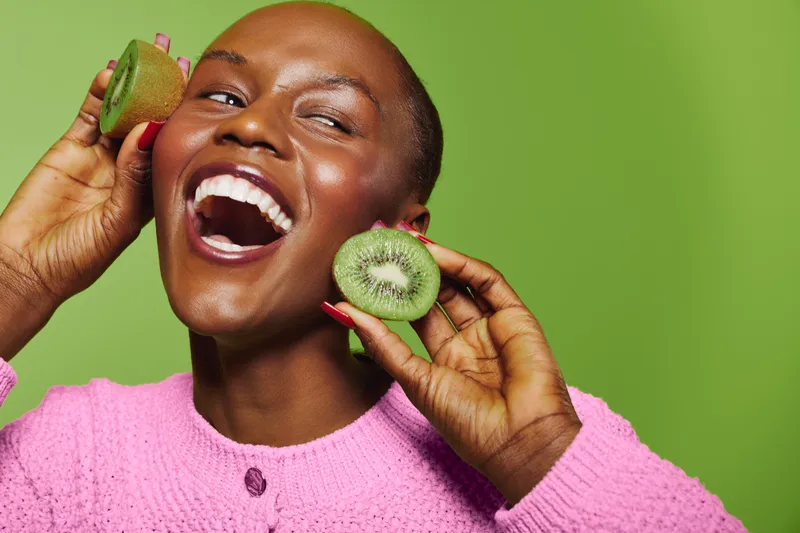 Happy African woman with dark skin holding kiwi slices near face smiles broadly against green background. Concept of healthy lifestyle, fresh fruit, natural beauty, and wellness.