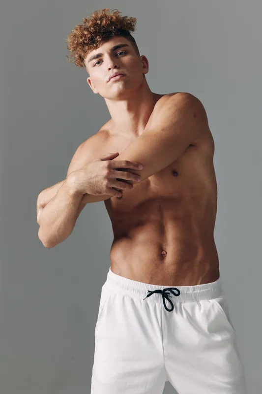 Young man with curly hair and muscular body posing shirtless in white shorts against gray background. Fitness, confidence and healthy lifestyle concept with toned skin and casual style.