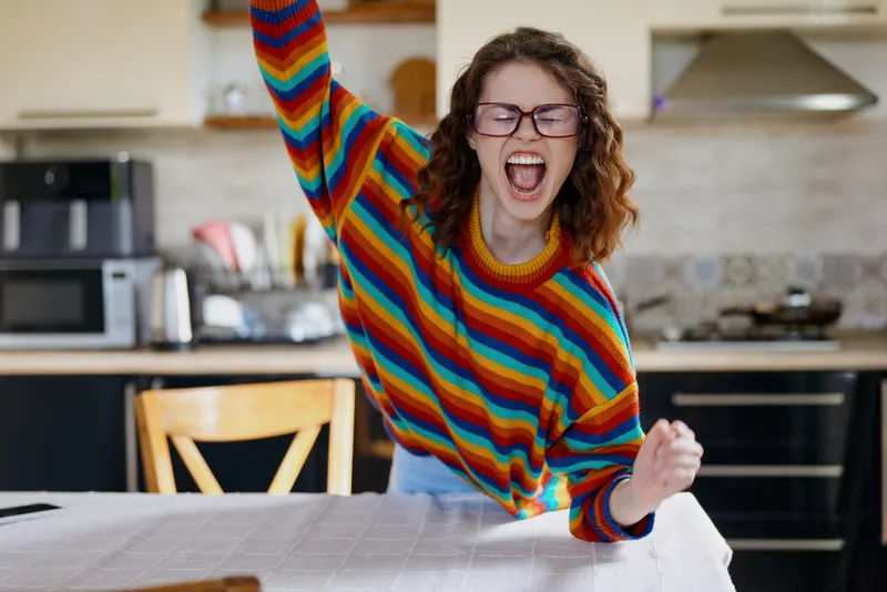 Joyful woman at home, standing by a table in a bright kitchen, laughing with arms raised, colorful striped sweater, daytime sunlight, vivid mood, authentic candid lifestyle moment