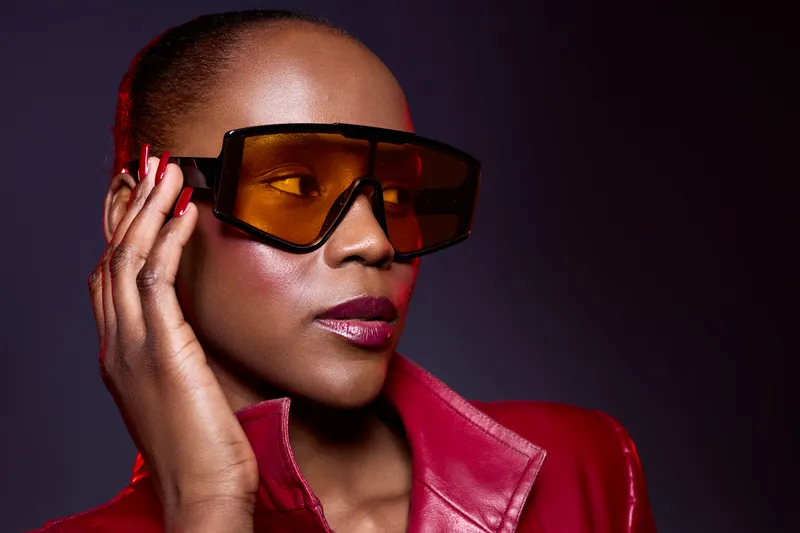Stylish African American woman with dark skin wearing large orange sunglasses and a red leather jacket poses confidently against a dark background. Fashion and attitude concept.