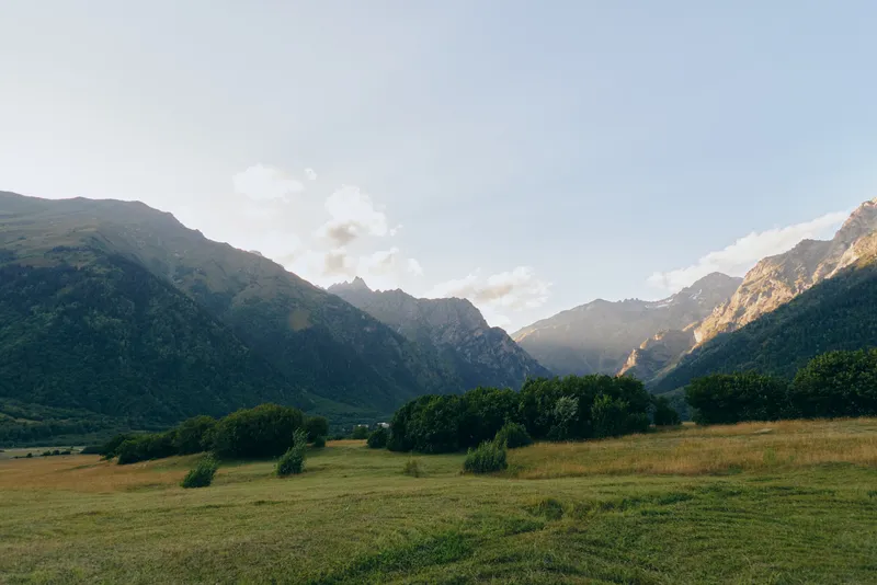 Serene mountain valley landscape featuring green meadow, scattered trees, and distant cliffs under a clear blue sky, suitable for nature and outdoor lifestyle imagery.