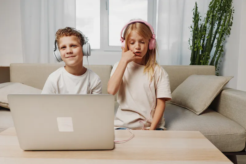 Two children wearing headphones use a laptop at home sitting on a couch in a bright living room. Concept for online learning, remote education, and digital communication.