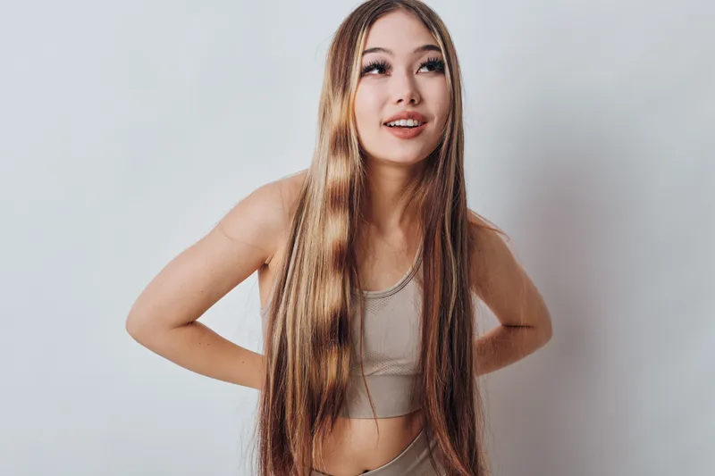 Young woman with long braided hair wearing a crop top and posing against gray studio background looking to the side with a smile and relaxed posture.