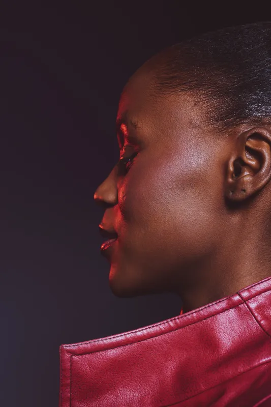 Profile of a dark skinned African woman with smooth skin wearing a red leather jacket. Studio portrait with dramatic lighting on a dark background. Concept of beauty and fashion.