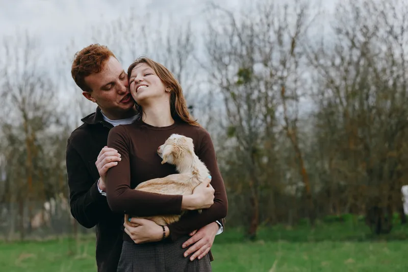 Lifestyle moment of a couple embracing outdoors with a small dog, warm affection and relaxed smiles, natural greens and bare trees in the background, candid peaceful atmosphere