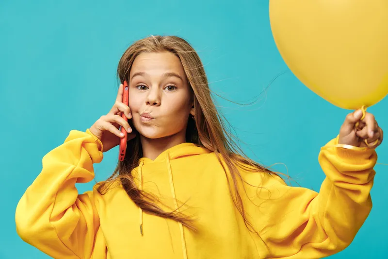 Girl in yellow hoodie talking on phone and holding yellow balloon with playful expression on blue background. Youth, communication, casual wear, lifestyle and colorful concept