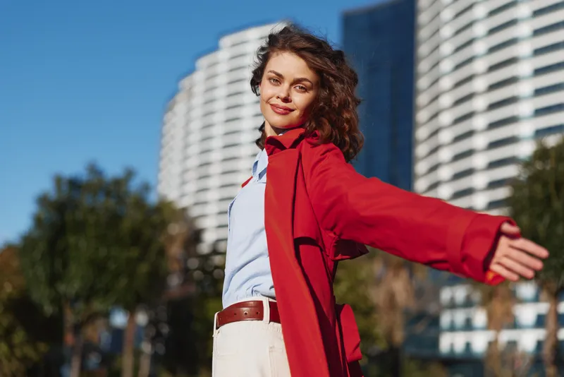 Young woman with curly hair wears red coat and white shirt while standing outdoors with arms outstretched and smiling confidently in urban setting.