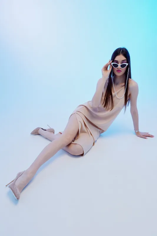 Woman beauty fashion model in beige dress and high heels posing on light background. She wears white sunglasses and pearl necklace. Studio shot with modern style and elegance.