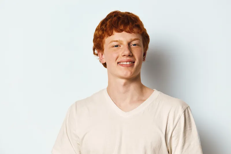 Young man with red hair and freckles wearing white t shirt smiling and looking at camera on plain white background. Casual portrait of confident happy male model.