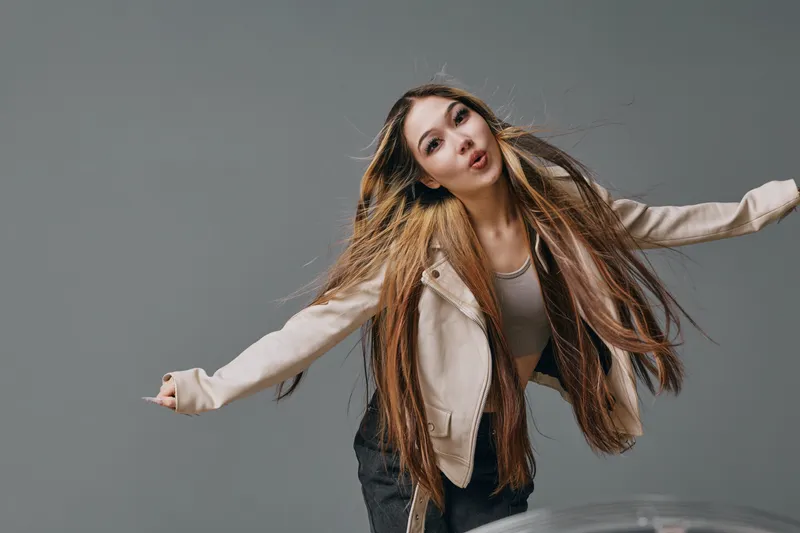 Young woman with long hair wearing casual jacket poses with arms extended against gray background in studio for portrait fashion beauty and lifestyle.