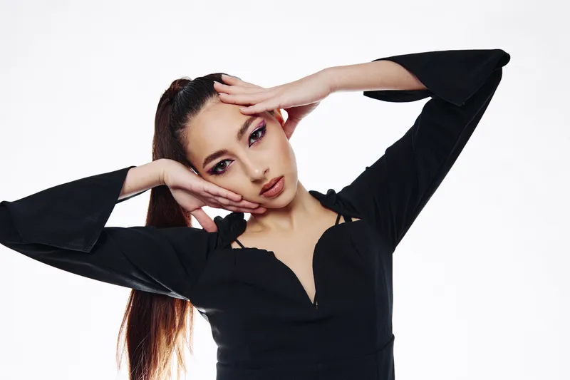 Young woman with flawless skin and dramatic makeup poses creatively with hands framing face against a white background. Fashion concept with focus on beauty and elegance.