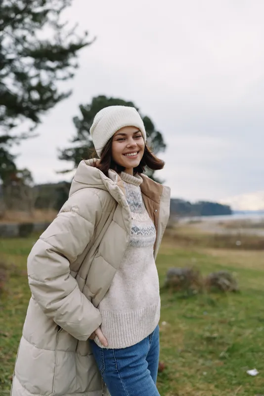 Woman outdoors wearing a beige puffer coat, knitted sweater and beanie, smiling in a breezy countryside setting with trees and grass, fashion lifestyle shot for cold weather apparel.