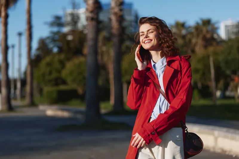Cheerful woman in a red coat stands outdoors on a sunny day, smiling warmly, posing confidently in a park with palm trees and clear blue sky