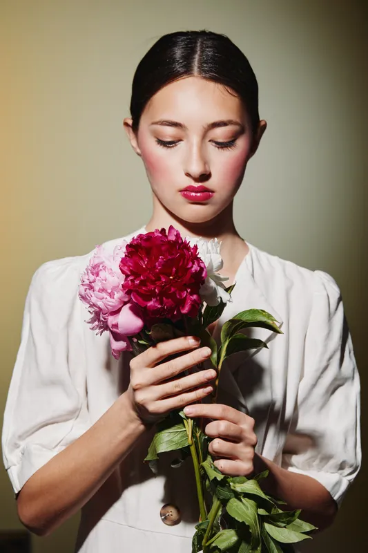 Young woman with pale skin and bold makeup holds a bouquet of colorful flowers close to her chest in natural light. Concept of beauty, fashion, creativity, and elegance.