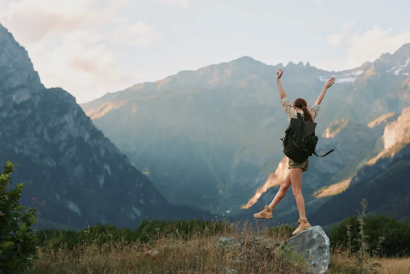 Young woman with backpack stands on a rocky ledge, arms raised in celebration, surrounded by rugged mountains and a clear blue sky, her casual outfit and excited expression conveying freedom and