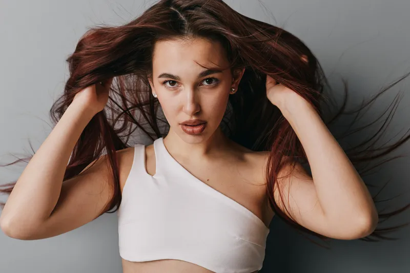 Young woman with long hair wearing white top posing indoors with hands in hair and serious facial expression against neutral background. Portrait of confident female model in casual style.