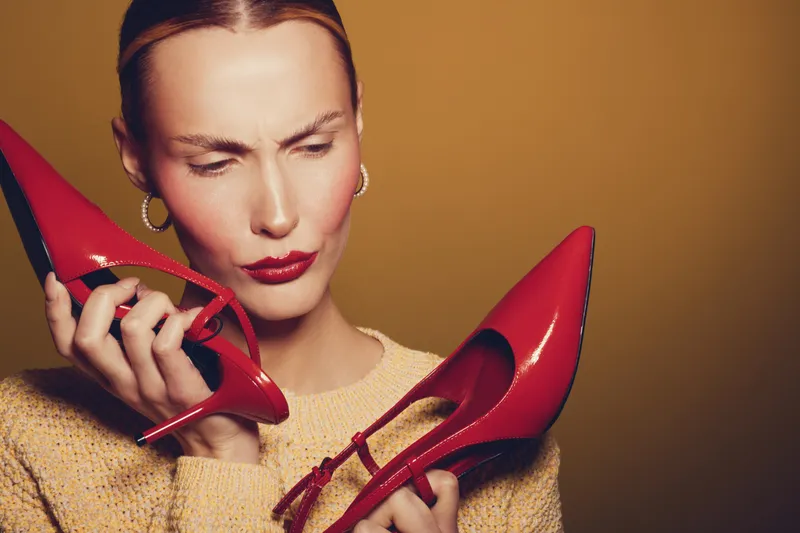 Young woman in yellow sweater holds red high heels and looks confused. Studio portrait on brown background. Concept for fashion choice, style, and footwear decision.