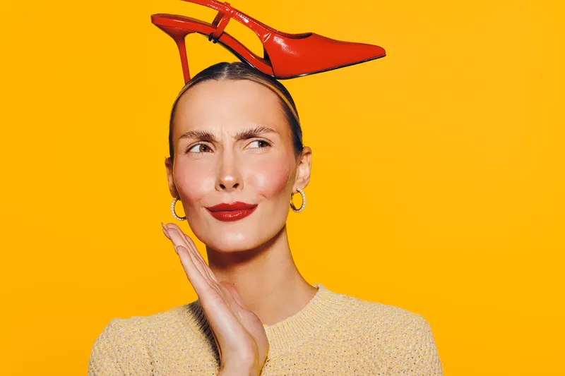 Young woman with red high heel shoe on head looks sideways with thoughtful expression on bright orange background. Concept for fashion, creativity, and playful style.