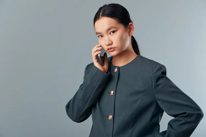Young woman in dark blazer with gold buttons talking on phone in studio with gray background. Businesswoman with serious expression standing with hand on hip and mobile device communication.