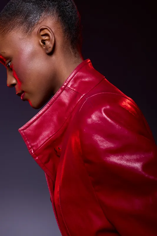 Profile of a young black woman wearing a shiny red leather jacket, against a dark background. The image highlights her smooth skin and elegant posture in studio lighting.