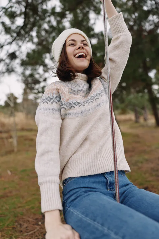Smiling woman in a cozy knit sweater and beanie enjoys a rope swing in an outdoor park, capturing carefree autumn moments and casual seasonal style.
