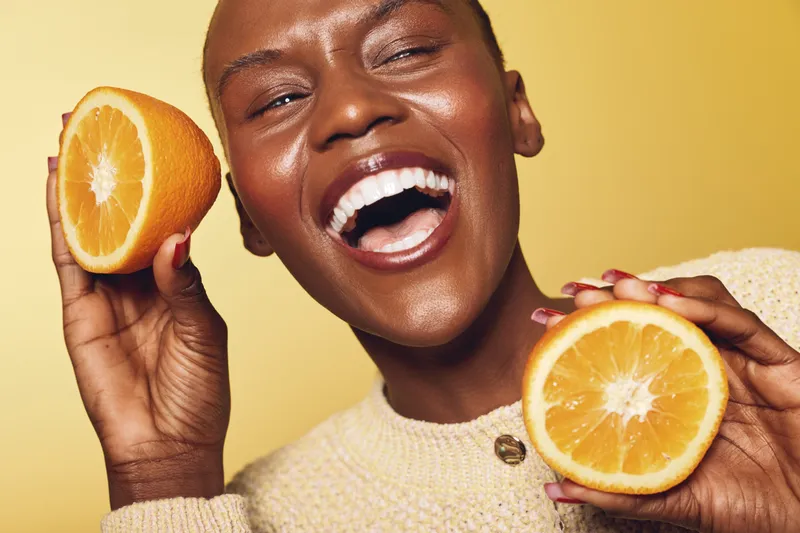 Happy dark skinned African woman with bright smile holds two halves of fresh orange fruit close to her face on yellow background. Concept of healthy lifestyle, fruit, vitamin and joy.